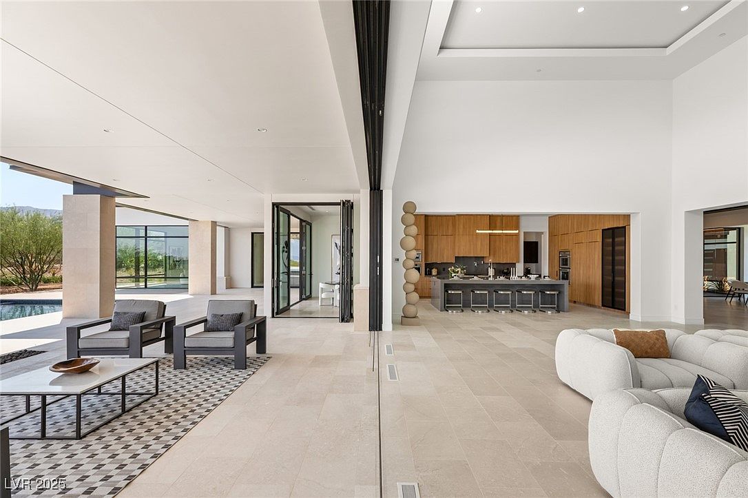 This is an interior shot of a modern living room that seamlessly blends indoor and outdoor spaces. The room features neutral tones, contemporary furniture, and large windows that offer views of the exterior. The open floor plan connects the living area to a modern kitchen with a large island and sleek cabinetry, creating a spacious and inviting atmosphere.