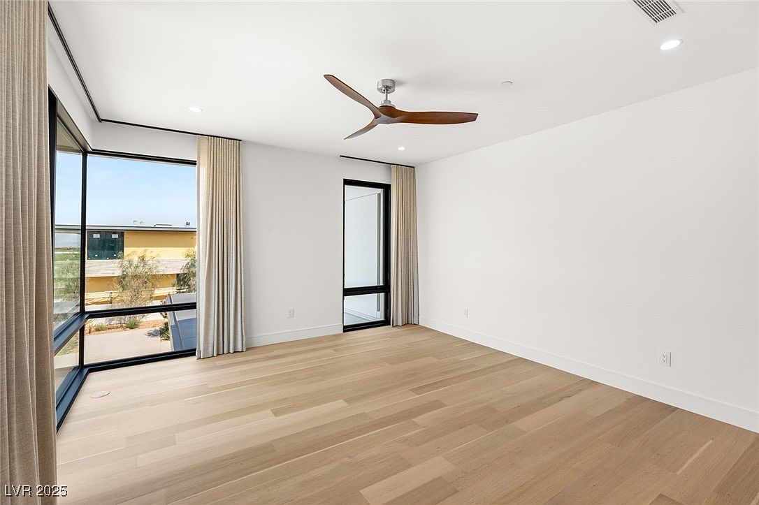 This is a bright and airy primary bedroom featuring light hardwood floors and white walls. Large windows with neutral-toned curtains provide ample natural light and views of the outside. A modern ceiling fan adds a touch of contemporary style to the room.