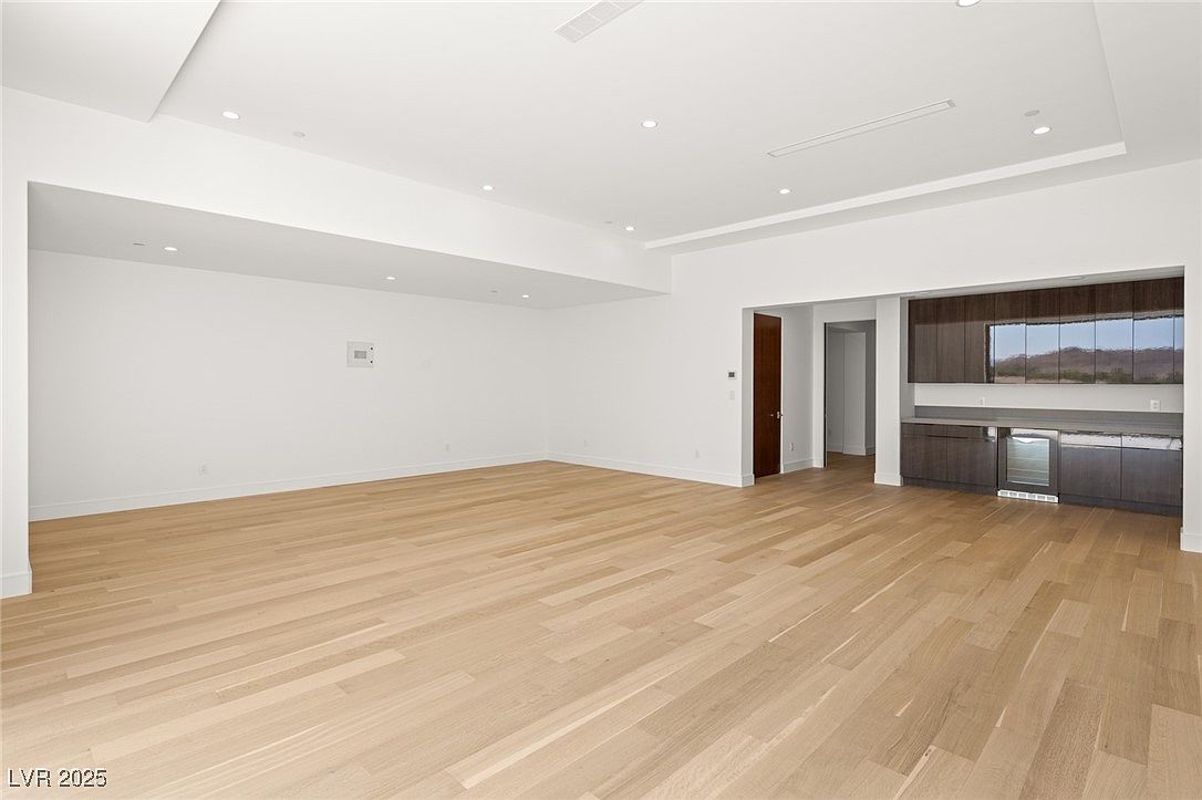 This is a spacious living room featuring light wood flooring and white walls, creating a bright and airy atmosphere. A built-in bar area with dark wood cabinetry and a beverage refrigerator adds a touch of sophistication. The room is well-lit with recessed lighting, and a doorway leads to another area of the house.