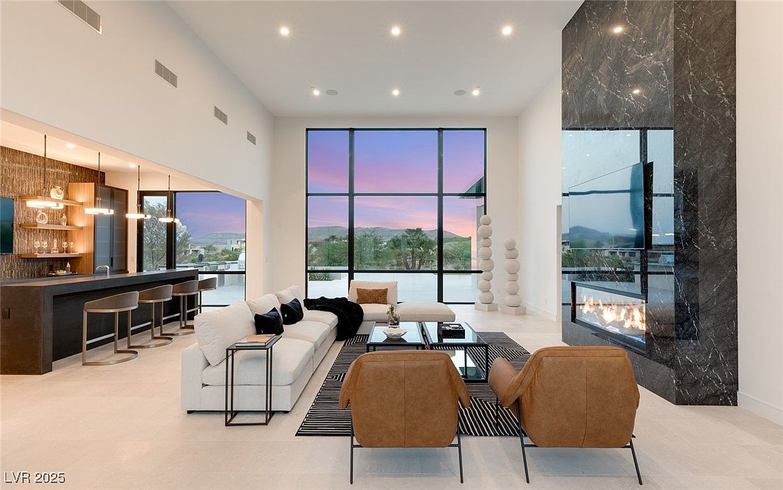 This is a spacious and modern living room featuring high ceilings, a large window with a view, and a striking marble fireplace. The room is furnished with a white sofa, tan leather chairs, and a glass coffee table, creating a luxurious and inviting atmosphere. The open floor plan seamlessly connects to a bar area, enhancing the sense of space and flow.