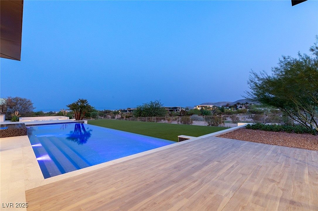 This image showcases a luxurious outdoor pool area at dusk, featuring a sleek, modern pool with illuminated steps and crystal-clear water reflecting the sky. The pool is surrounded by a spacious wooden deck and manicured lawn, with desert landscaping and distant mountain views adding to the serene ambiance. The scene evokes a sense of high-end living and relaxation.