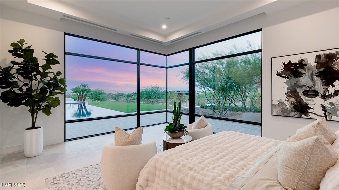 This is a luxurious primary bedroom featuring floor-to-ceiling windows with views of the pool and landscape. The room is decorated in neutral tones with a plush bed, comfortable seating, and modern art. The overall impression is one of serenity and high-end comfort.