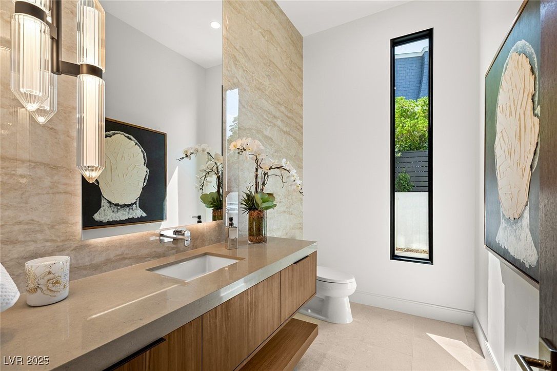 This is a modern bathroom featuring a floating vanity with a light wood finish and a beige countertop. A large mirror reflects the vanity area, and a narrow window provides natural light. The walls are a combination of white and beige marble, and the overall style is minimalist and elegant.