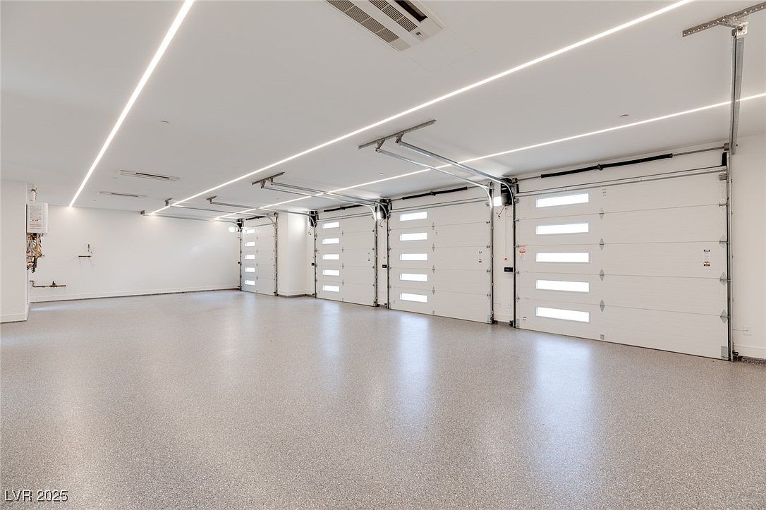 The image showcases a spacious and well-lit garage with multiple white garage doors featuring horizontal windows. The floor is a speckled gray epoxy, and the ceiling has modern linear lighting. The overall impression is clean, organized, and high-end, suggesting a luxury property.