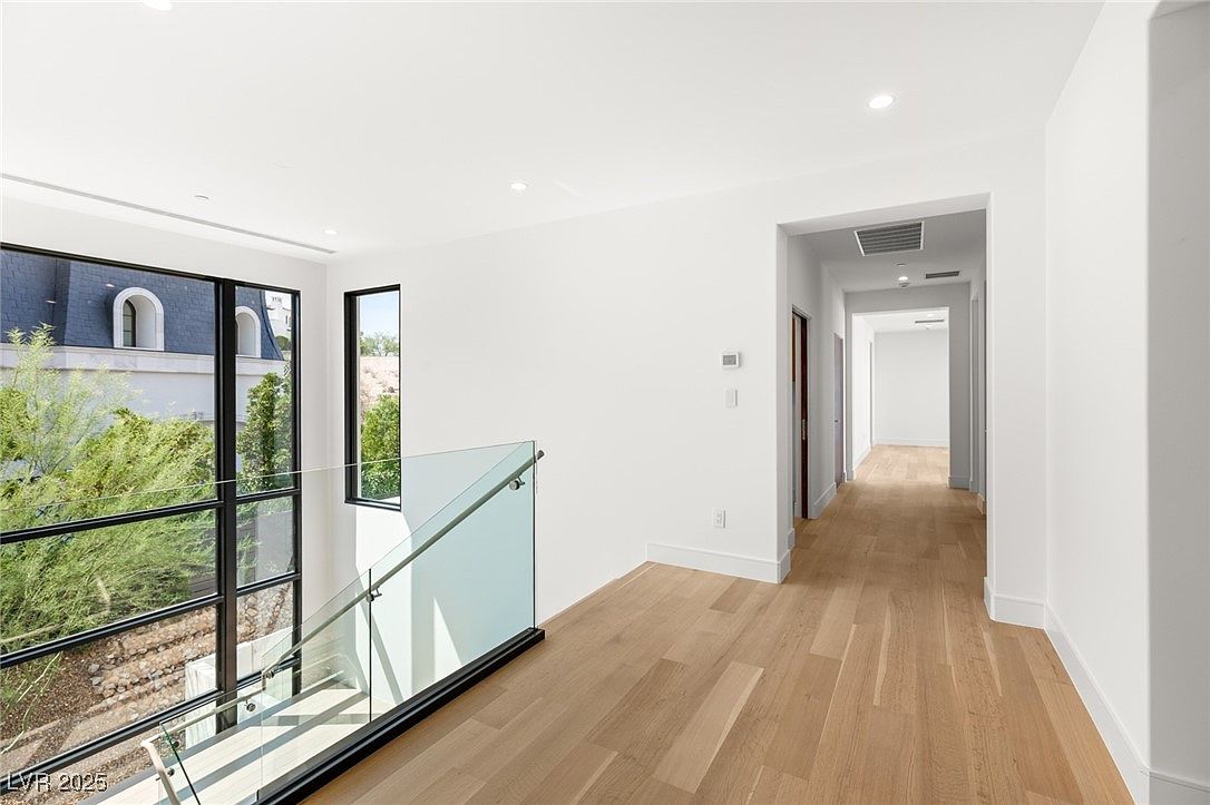 This interior shot showcases a bright and modern hallway with light hardwood flooring and white walls. A glass railing provides a sleek barrier along the staircase, offering a view through large windows to the exterior. The hallway leads to other rooms, suggesting a spacious and well-lit home layout.