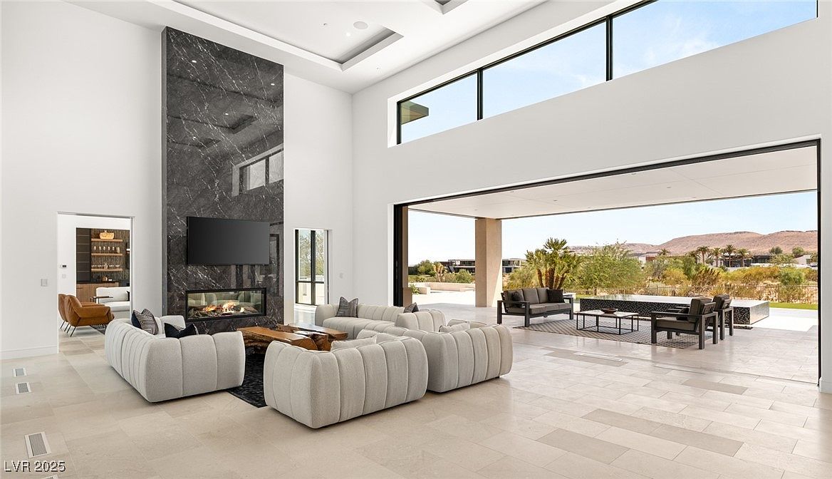 This is a spacious, modern living room with high ceilings and an open floor plan that seamlessly connects to an outdoor patio. The room features a striking marble fireplace, a large television, and comfortable, modular seating. The expansive sliding glass doors offer panoramic views of the landscape, creating an indoor-outdoor living experience.
