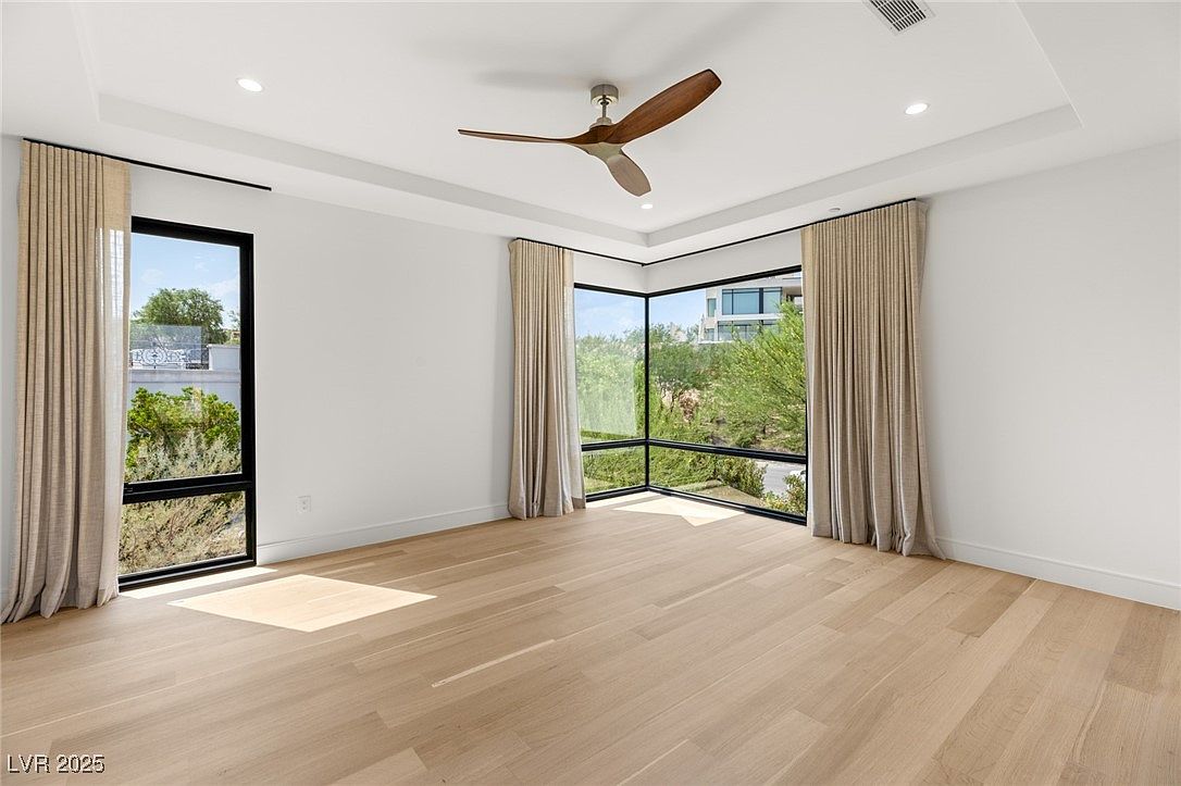 This is a bright and spacious primary bedroom featuring hardwood floors and large windows that offer ample natural light and views of the surrounding landscape. The room is minimally furnished, highlighting the clean lines and modern design. A ceiling fan adds to the comfort of the space.