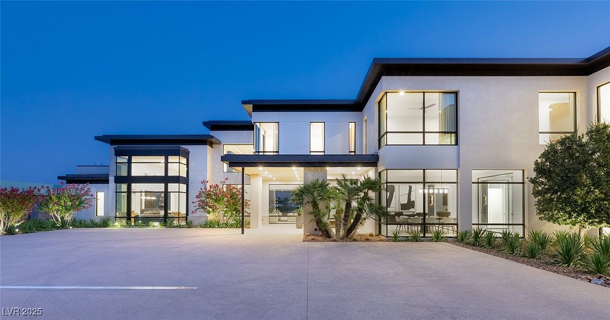 This is a front exterior view of a modern, multi-story home at dusk. The house features a flat roof, large windows with black frames, and a light-colored facade. The driveway is spacious and leads to a covered entryway, with manicured landscaping and palm trees adding to the property's curb appeal.
