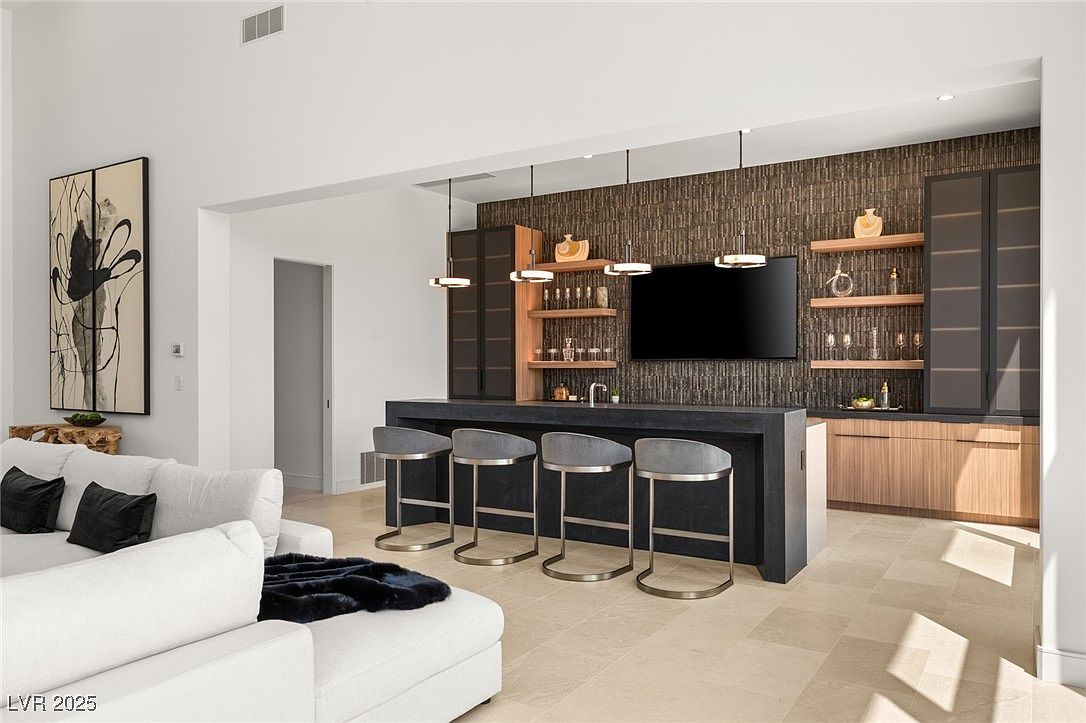 This interior shot showcases a modern living room with a stylish bar area. The bar features a dark countertop, seating for four, and a backdrop with a large television and shelving. The living room area includes a white sectional sofa with dark pillows and a throw, creating a comfortable and inviting space.