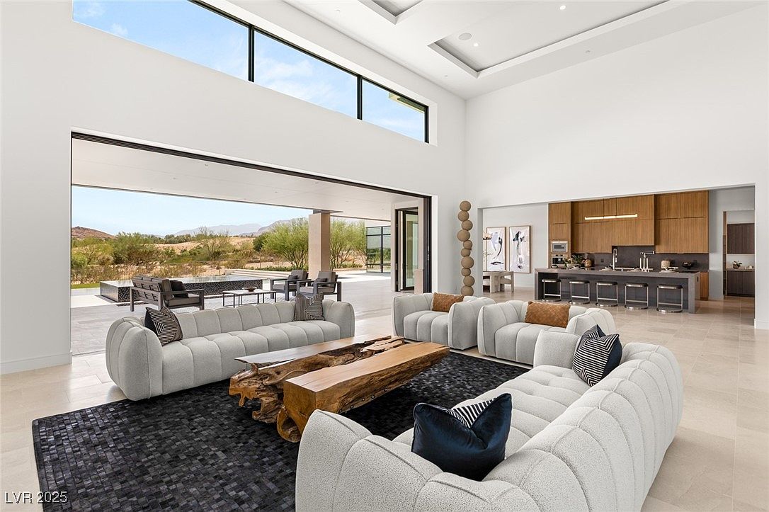 This is a spacious and luxurious living room featuring high ceilings and an open floor plan that seamlessly connects to the outdoors. The room is furnished with modern, curved sofas, a unique wooden coffee table, and a stylish rug. Large sliding glass doors provide access to an outdoor patio and offer stunning views of the surrounding landscape, while the kitchen is visible in the background.