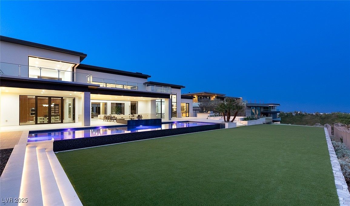 This is a rear view of a modern luxury home featuring a sleek design with large glass windows and doors. A swimming pool with illuminated water and a well-manicured lawn extend from the house, creating an inviting outdoor space. The perspective is wide, showcasing the scale of the property and its integration with the landscape.