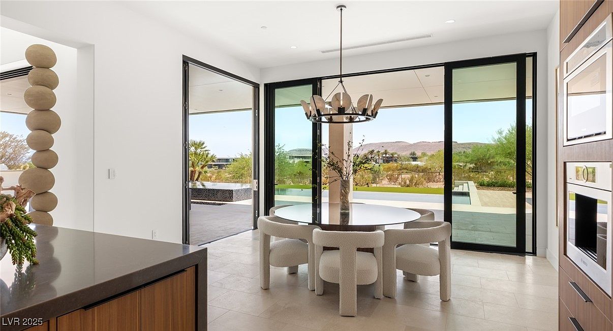 This is an interior shot of a modern dining room featuring a round glass table with unique, upholstered chairs. Large sliding glass doors offer an expansive view of the outdoor landscape, seamlessly blending indoor and outdoor living. A stylish chandelier hangs above the table, adding a touch of elegance to the minimalist design.