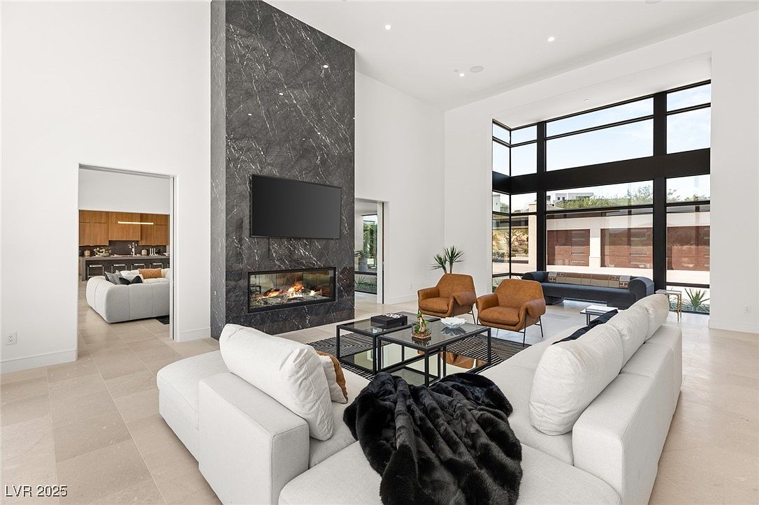 This is an interior shot of a modern living room featuring a sleek fireplace with a large television mounted above it. The room is furnished with a white sectional sofa, brown leather armchairs, and a glass coffee table. Large windows provide ample natural light and a view of the outdoors, enhancing the spacious and airy feel of the room.