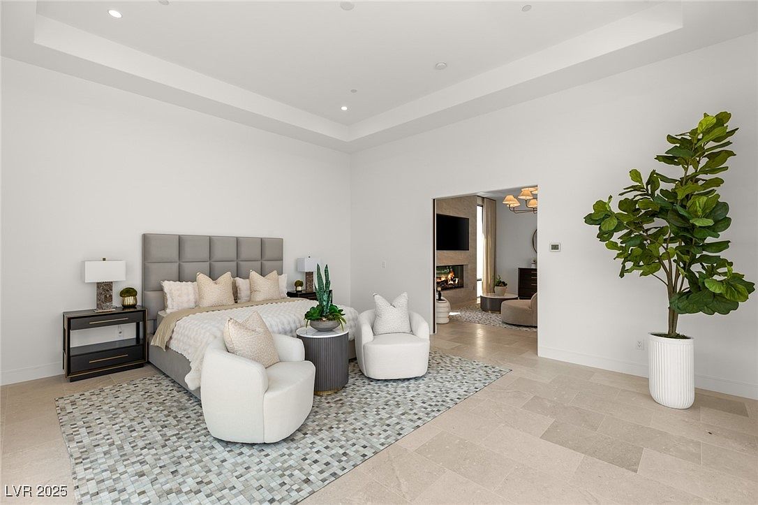This is a bright and airy primary bedroom featuring a neutral color palette. The room includes a large bed with a tufted headboard, two armchairs, and a side table with a lamp. A doorway leads to another room, and a large potted plant adds a touch of greenery.