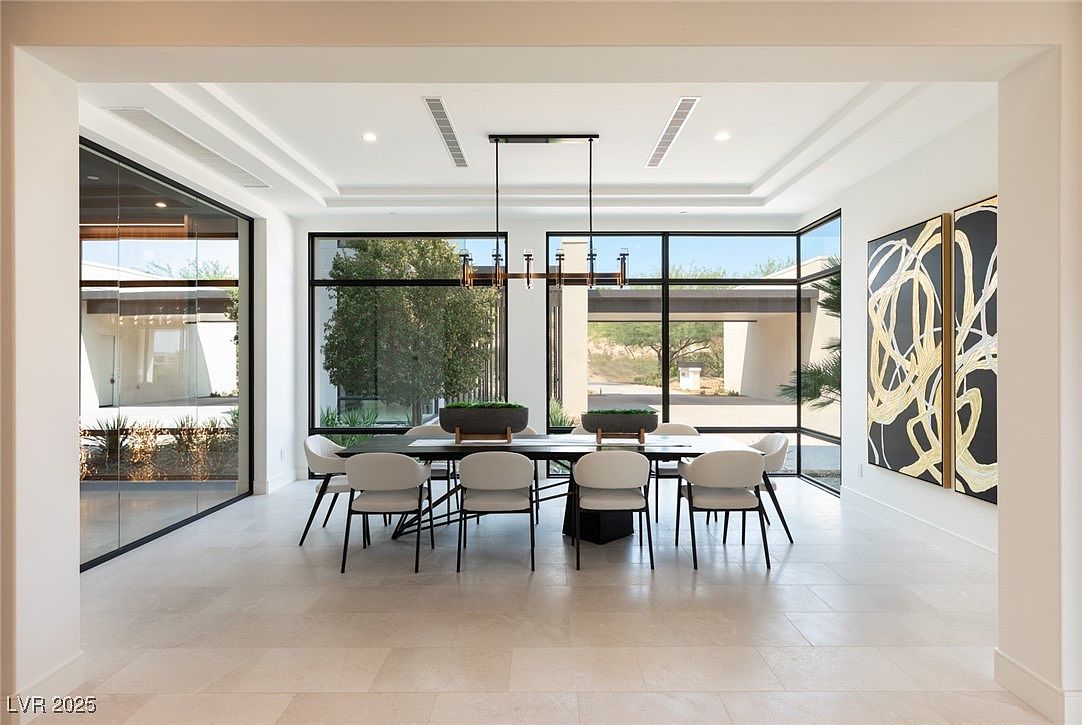 This is an interior shot of a modern dining room. The room features a long dining table with seating for eight, a contemporary chandelier, and large windows offering an abundance of natural light and views of the outdoors. The walls are painted in a neutral tone, and there are modern art pieces on the wall, contributing to the room's sophisticated and minimalist aesthetic.
