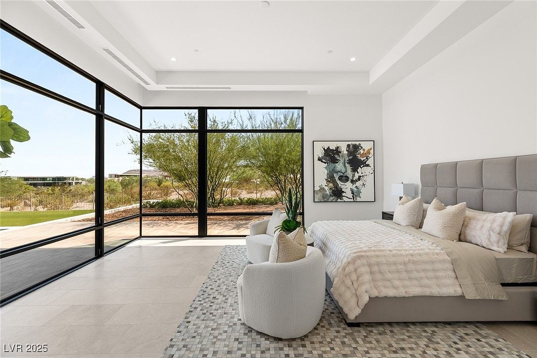 This is a bright and spacious primary bedroom featuring floor-to-ceiling windows that offer an expansive view of the outdoors. The room is decorated in a modern style with a neutral color palette, including a large upholstered headboard, plush bedding, and a contemporary area rug. Two armchairs create a cozy seating area near the windows.