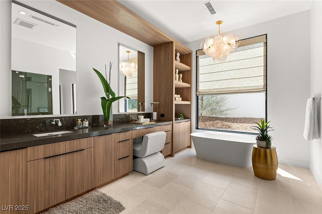 This is a luxurious primary bathroom featuring a long vanity with wood cabinetry and a dark countertop. A large mirror reflects the space, and a modern chandelier hangs above a freestanding bathtub near a window with blinds. The room is spacious and well-lit, creating a spa-like atmosphere.