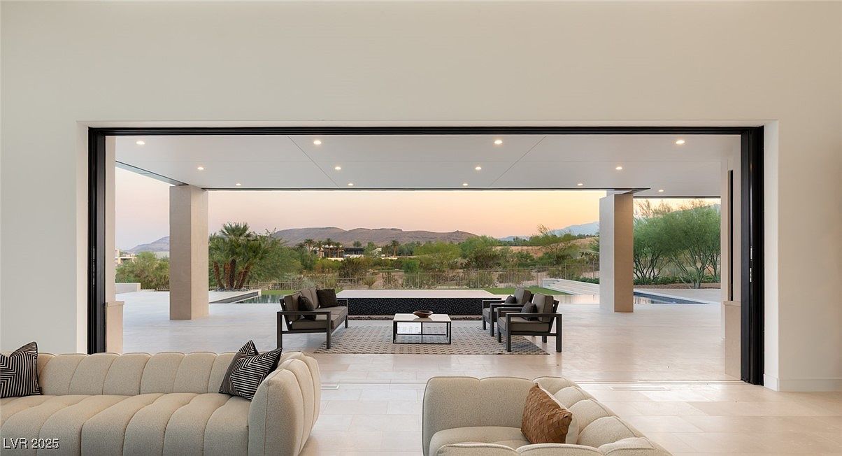 This image showcases a luxurious living room that seamlessly blends indoor and outdoor spaces. Large sliding glass doors open to a patio area with outdoor seating, a water feature, and views of distant mountains. The interior features modern furniture, neutral color palettes, and clean lines, creating an elegant and inviting atmosphere.