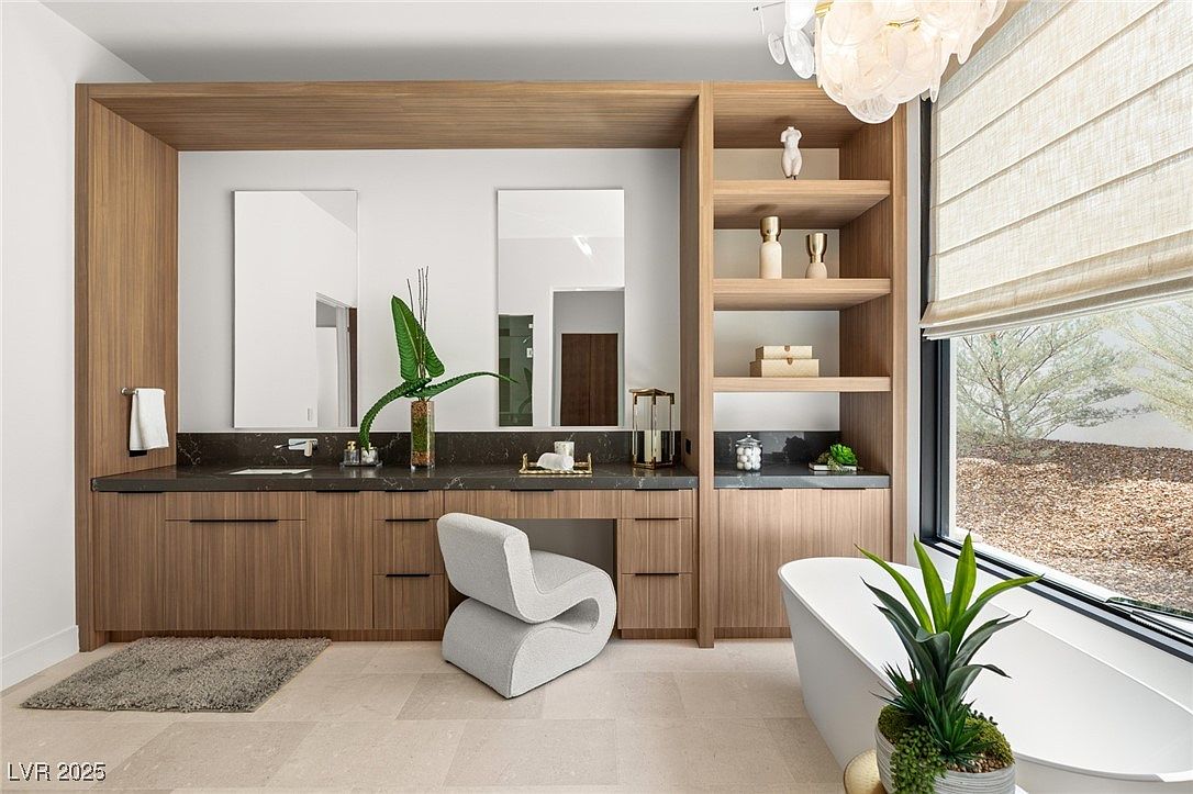 This is a luxurious primary bathroom featuring a modern vanity with dual sinks and dark countertops. The space is well-lit with natural light from a large window, complemented by stylish shelving and a unique chair. A freestanding bathtub adds to the spa-like atmosphere, enhanced by a decorative plant.