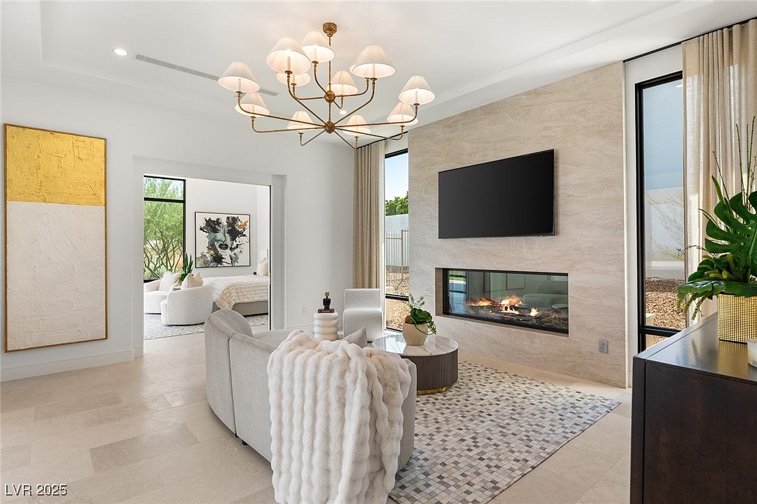 This is an interior shot of a modern living room featuring a linear fireplace and a large television mounted above it. The room is decorated in neutral tones with a light gray sofa, a round coffee table, and a patterned rug. A doorway leads to another room, and a large piece of artwork hangs on the wall, adding a touch of color.