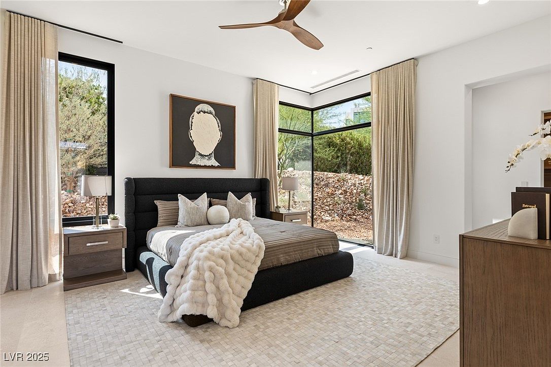 This is a primary bedroom featuring a large bed with a dark headboard and neutral bedding, complemented by a textured white throw. The room has large windows with natural light and neutral-toned curtains, along with modern artwork and a ceiling fan. The flooring is a light-colored tile or stone, and the overall style is contemporary and serene.