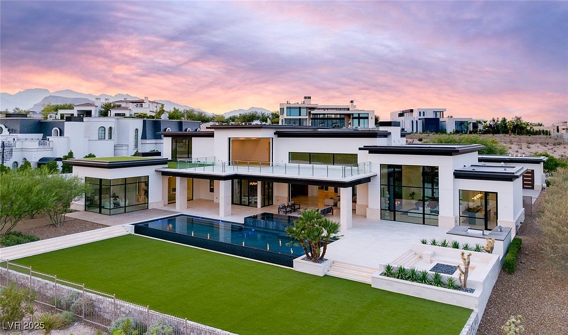 This image showcases the rear exterior of a modern luxury home, featuring a sleek design with clean lines, a flat roof, and expansive glass windows and doors. A rectangular swimming pool is centrally located, surrounded by a well-manicured lawn and a spacious patio area perfect for outdoor entertaining. The view includes other modern homes in the background and a picturesque sky at dusk.