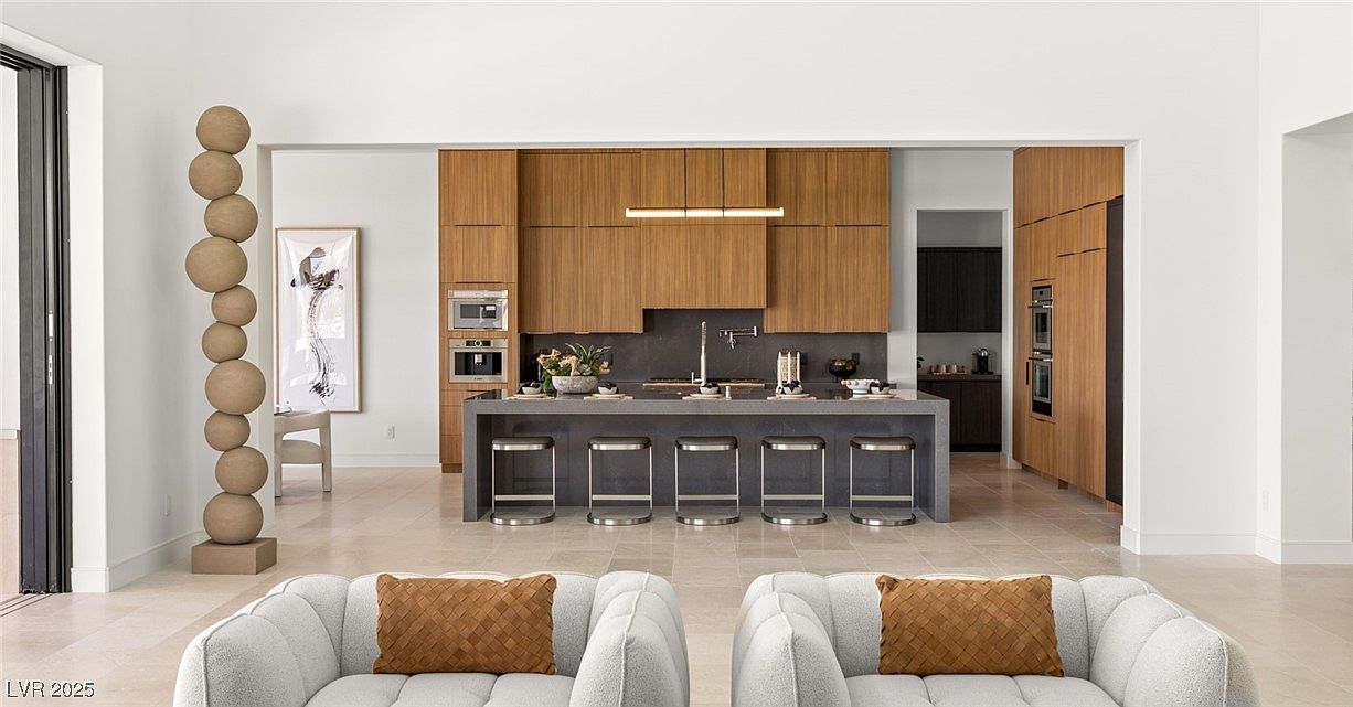 This interior shot showcases a modern kitchen with sleek, wood-paneled cabinetry and a gray countertop island featuring bar seating. The open-concept design flows into an adjacent space, enhancing the sense of spaciousness. The foreground includes a glimpse of stylish living room furniture, creating a cohesive and inviting atmosphere.