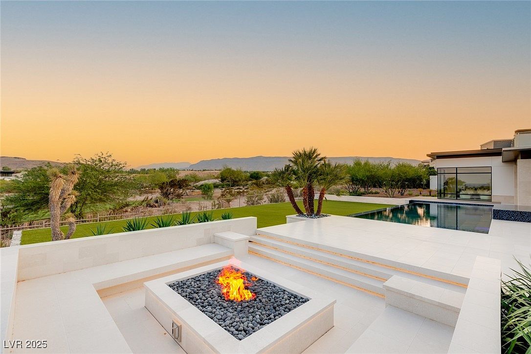 This image showcases a luxurious outdoor patio area featuring a modern fire pit surrounded by sleek, white seating. The patio extends to a pool area, with well-manicured landscaping and a scenic mountain view in the background. The overall impression is one of sophisticated outdoor living and relaxation.