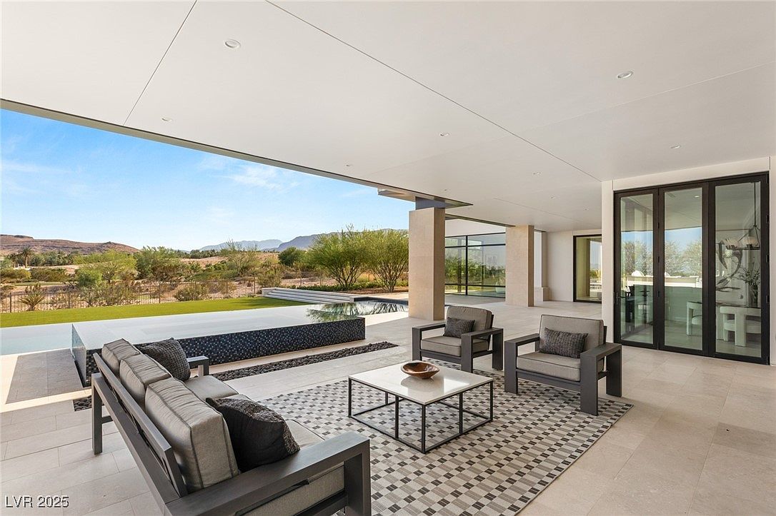 This image showcases a luxurious outdoor patio area, featuring modern furniture including a sofa, two armchairs, and a coffee table arranged on a patterned rug. The patio is covered by an extended roof, providing shade and creating an indoor-outdoor living space. Beyond the patio, there's a view of a pool, landscaping, and distant mountains, enhancing the property's appeal.