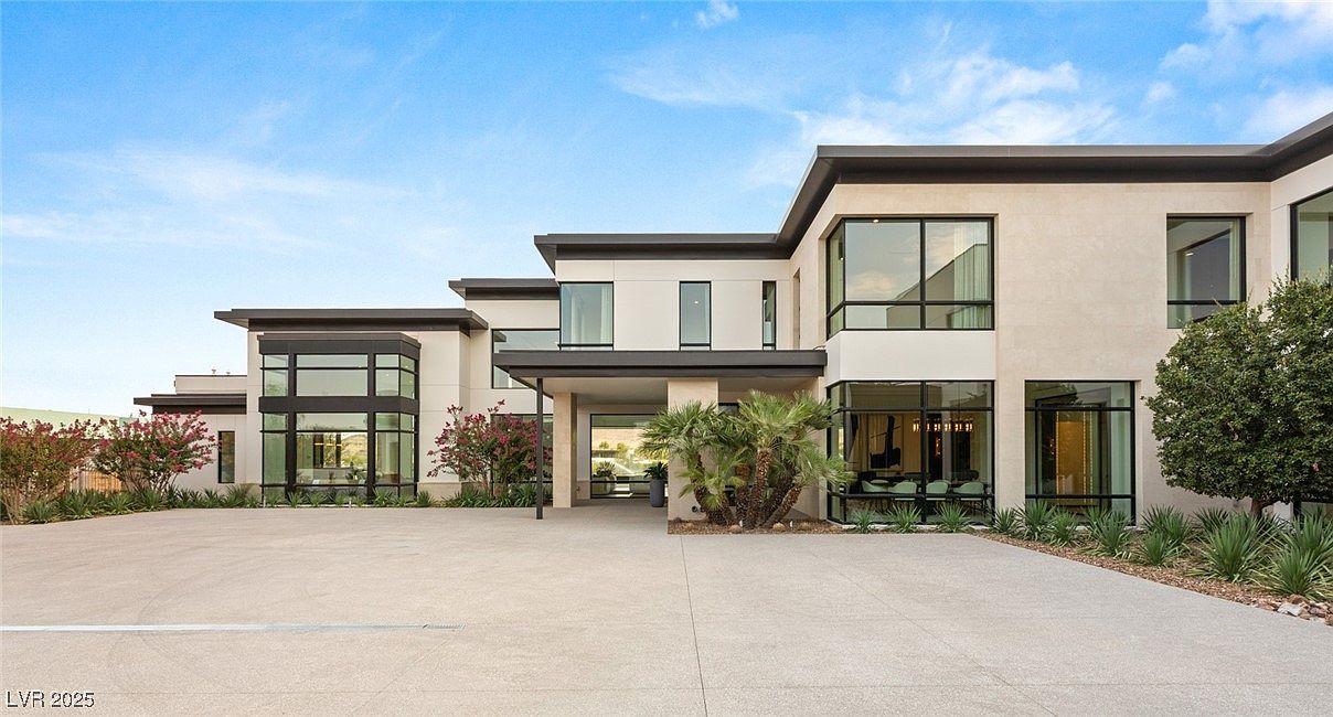 This is a front exterior view of a modern, multi-story home. The house features a flat roof, large windows with dark frames, and a light-colored facade. A spacious driveway leads up to the entrance, which is framed by landscaping including trees and shrubs, creating a welcoming and luxurious curb appeal.