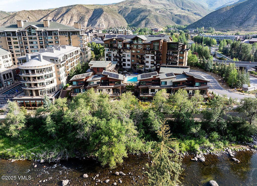 This aerial view showcases a luxurious residential complex nestled in a mountainous landscape. The architecture features a blend of modern and rustic elements, with multiple buildings, including condos and townhouses, surrounding a central pool area. Lush greenery lines the riverbank in the foreground, adding to the serene and upscale ambiance of the property.
