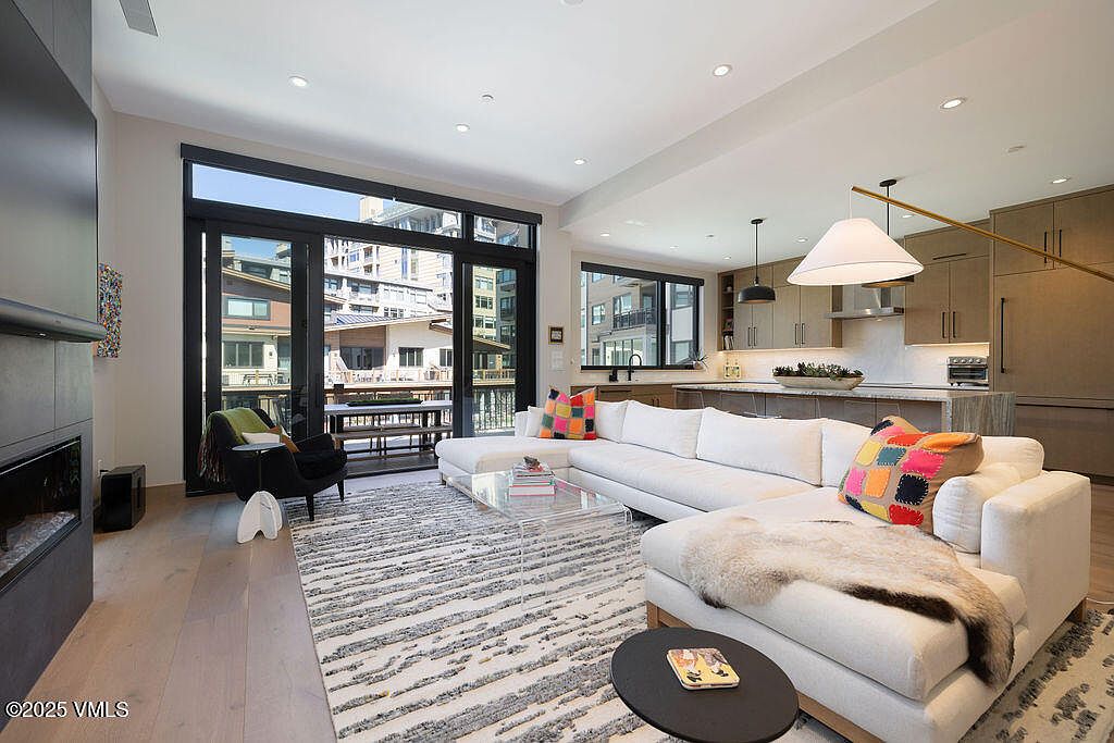 This is an interior shot of a modern living room featuring a large white sectional sofa with colorful pillows and a fur throw. The room has a contemporary rug, a clear coffee table, and a black accent chair. Large windows offer a view of the exterior, and the open floor plan seamlessly connects to the kitchen area, creating a spacious and inviting atmosphere.