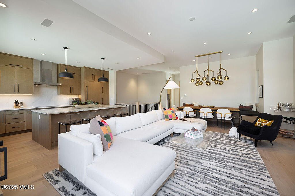 This is an interior shot of a modern living room that seamlessly blends with the kitchen and dining area. The living room features a large white sectional sofa, a patterned area rug, and a black accent chair. The kitchen boasts sleek cabinetry and a spacious island, while the dining area is illuminated by a unique chandelier, creating an open and inviting atmosphere.