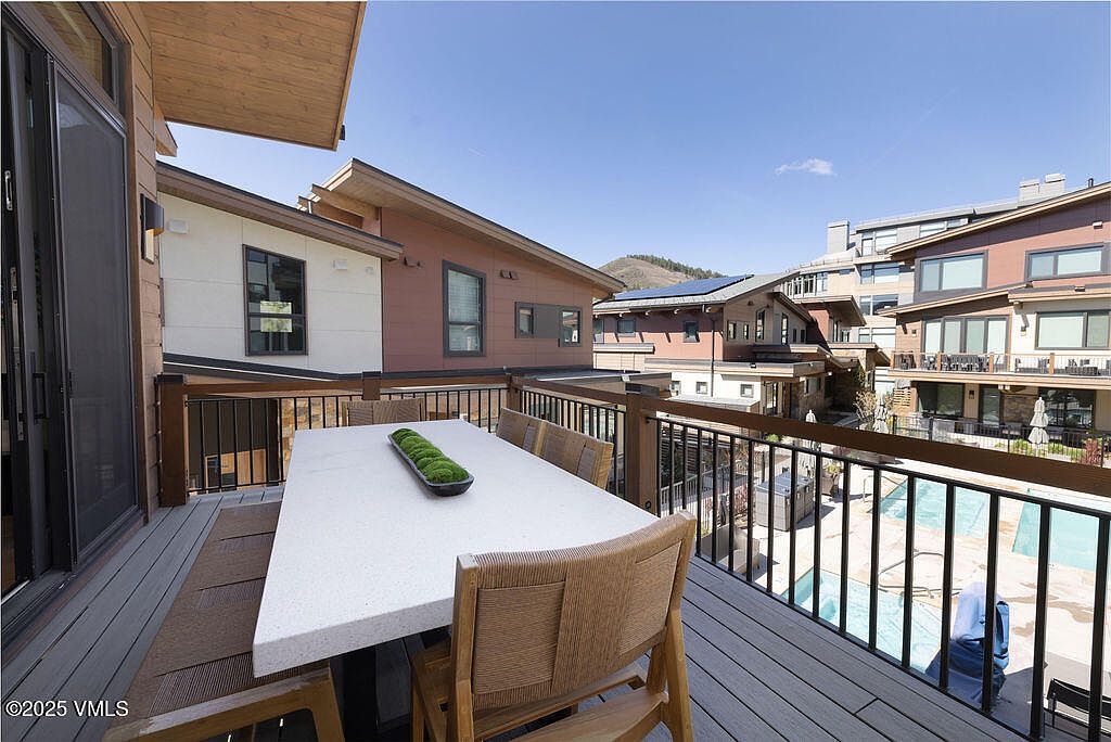 This image showcases a well-appointed patio or balcony area, featuring a modern outdoor dining set with a white table and woven chairs. The deck is constructed with composite decking, and a black metal railing provides safety and a clear view of the surrounding buildings and a glimpse of a pool area below. The setting suggests a luxurious outdoor living space perfect for entertaining or relaxing.