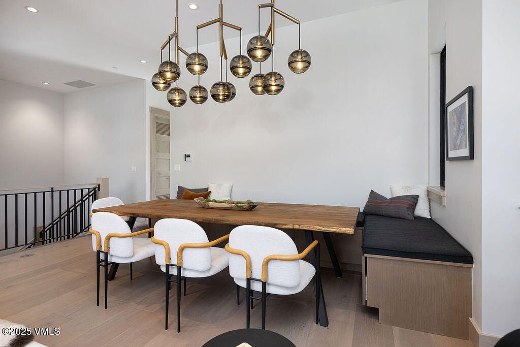 This is an interior shot of a dining room featuring a long wooden table with modern chairs and a built-in bench seating area. An eye-catching chandelier hangs above the table, providing ambient lighting. The room has a clean, contemporary aesthetic with white walls and light wood flooring, creating a bright and inviting space.