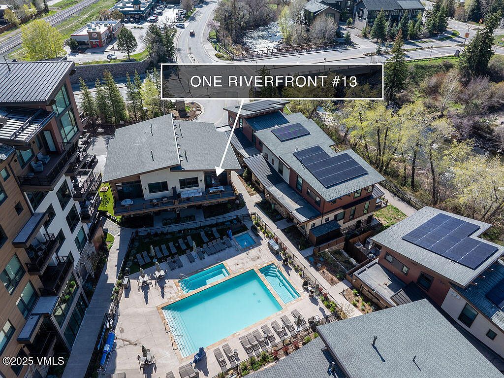 This aerial shot showcases a multi-unit residential complex featuring modern architecture, a central swimming pool, and well-maintained landscaping. The buildings have a mix of flat and pitched roofs, some with solar panels, and balconies offering outdoor living spaces. The pool area is furnished with lounge chairs, creating an inviting recreational space for residents.