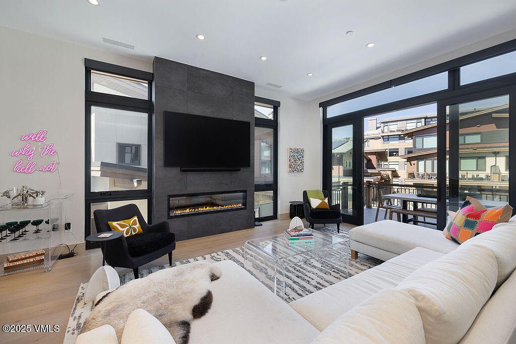 This is a bright and modern living room featuring a sleek fireplace with a large television mounted above. The room is furnished with a comfortable white sectional sofa, a clear acrylic coffee table, and stylish armchairs. Large windows and sliding glass doors provide ample natural light and access to an outdoor balcony, enhancing the indoor-outdoor living experience.