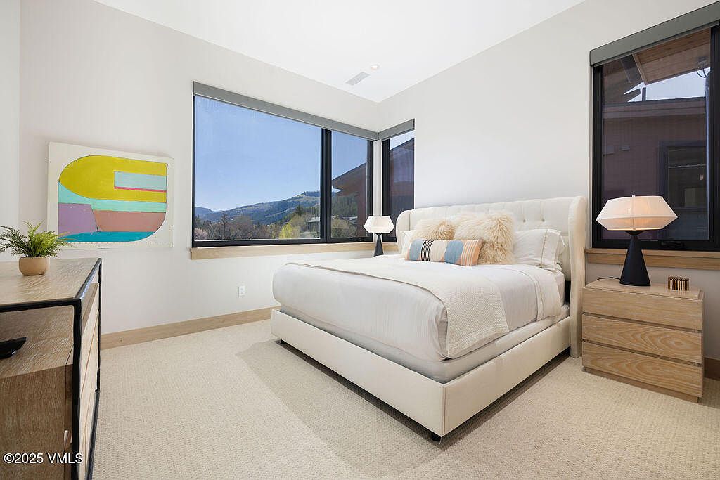 This is a bright and airy bedroom featuring a large window with mountain views. The room is furnished with a white upholstered bed, a light wood nightstand, and a modern art piece on the wall. The neutral color palette and plush carpeting create a serene and inviting atmosphere.
