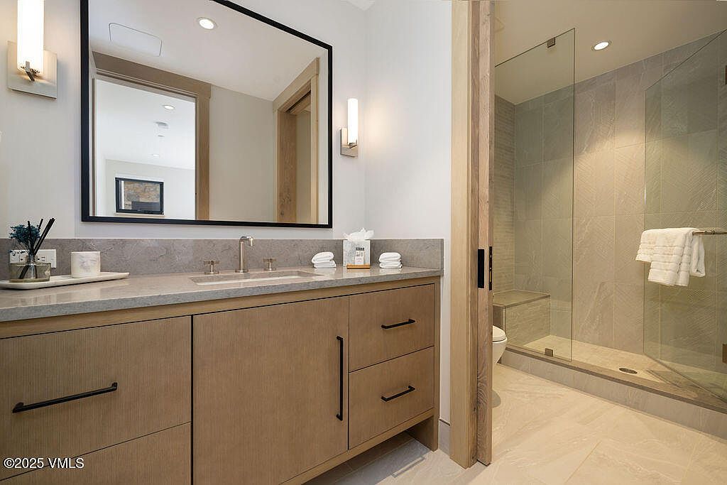 This is a primary bathroom featuring a modern vanity with a light wood finish, a gray countertop, and black hardware. A large mirror with a black frame hangs above the vanity, and sconces provide soft lighting. The bathroom includes a glass-enclosed shower with neutral-toned tiles and a built-in bench, creating a spa-like atmosphere.
