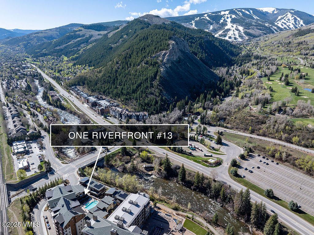 This aerial shot showcases the One Riverfront #13 property, highlighting its location near a river and a mountain range with ski slopes. The building features a pool and is surrounded by lush greenery and a parking area. The image emphasizes the property's scenic setting and proximity to both natural beauty and recreational opportunities.