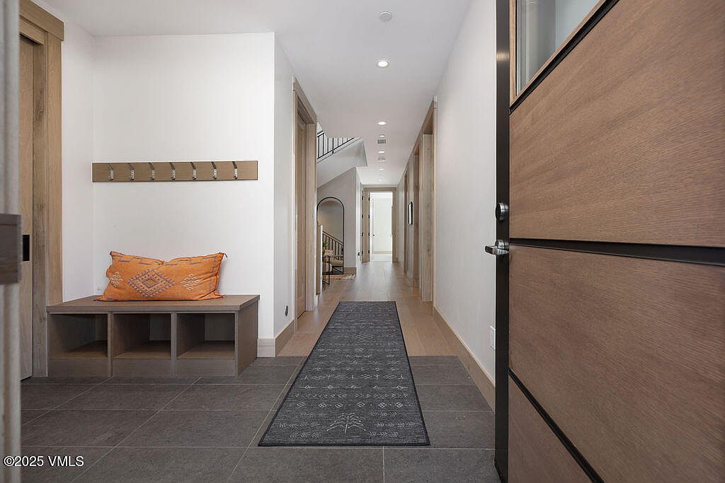 This interior shot showcases a modern hallway with a blend of textures and neutral tones. The entryway features a built-in bench with storage, topped with an orange pillow, and a coat rack above. A long runner rug leads down the hallway, drawing the eye towards the staircase and other rooms, while a contemporary wooden door is partially visible on the right.