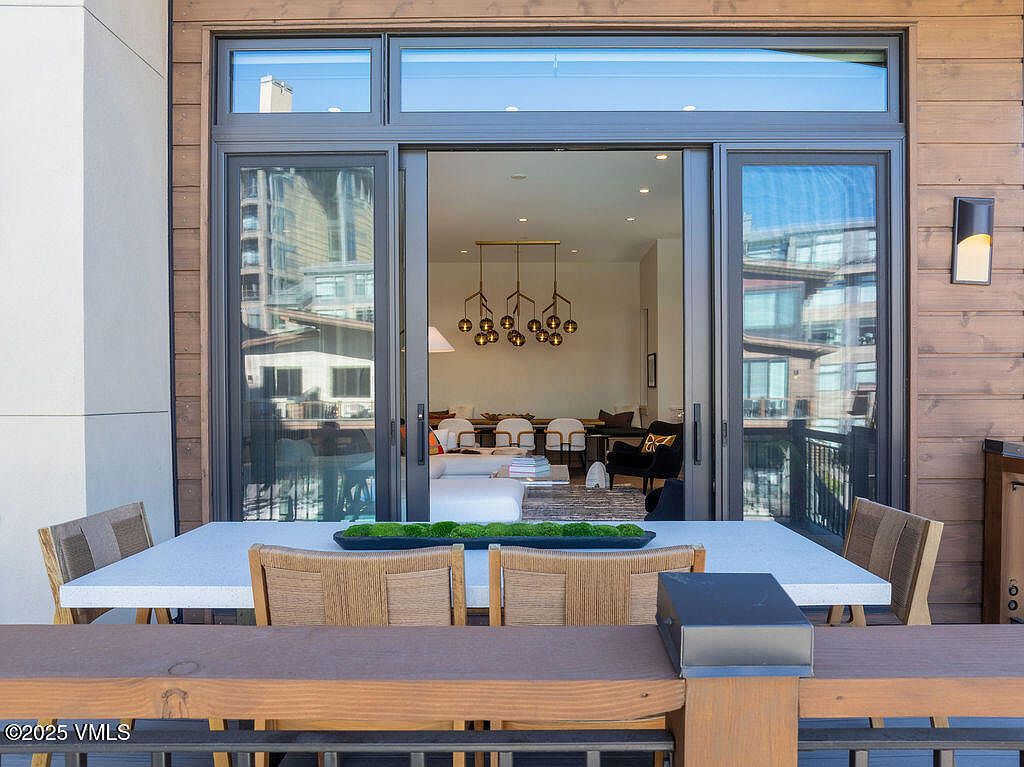 This image showcases a stylish patio or balcony area, featuring a modern outdoor dining set with a white speckled table and woven chairs. The space is connected to the interior via large sliding glass doors, offering a seamless transition between indoor and outdoor living. The view through the doors reveals a contemporary living space with a unique chandelier, suggesting a luxurious and inviting atmosphere.