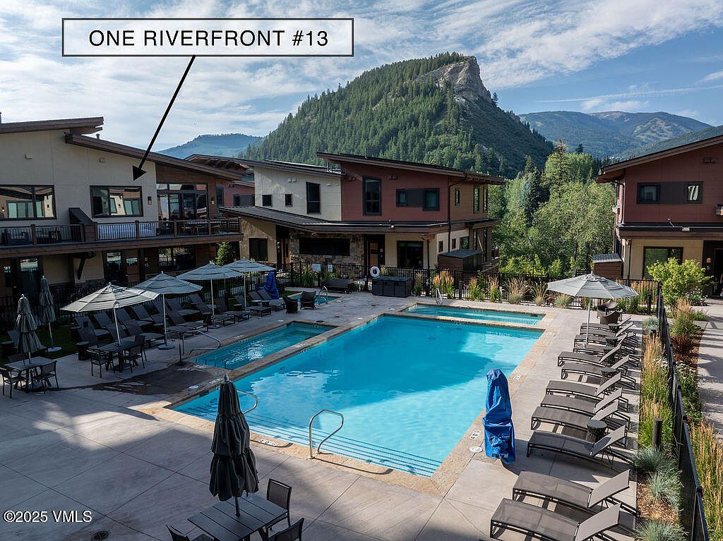 This image showcases a well-maintained outdoor pool and spa area, surrounded by lounge chairs and umbrellas, suggesting a relaxing and luxurious amenity. The architecture of the adjacent buildings is modern with a mountain backdrop, enhancing the appeal of the property. The pool's clear water and inviting layout create a desirable recreational space.