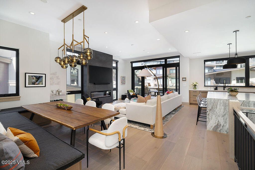 This is an interior shot of a modern living and dining area. The space features a dining table with bench seating, a contemporary chandelier, a sleek fireplace with a mounted TV, and a comfortable-looking sofa. Large windows and sliding glass doors provide ample natural light and a view of the exterior.