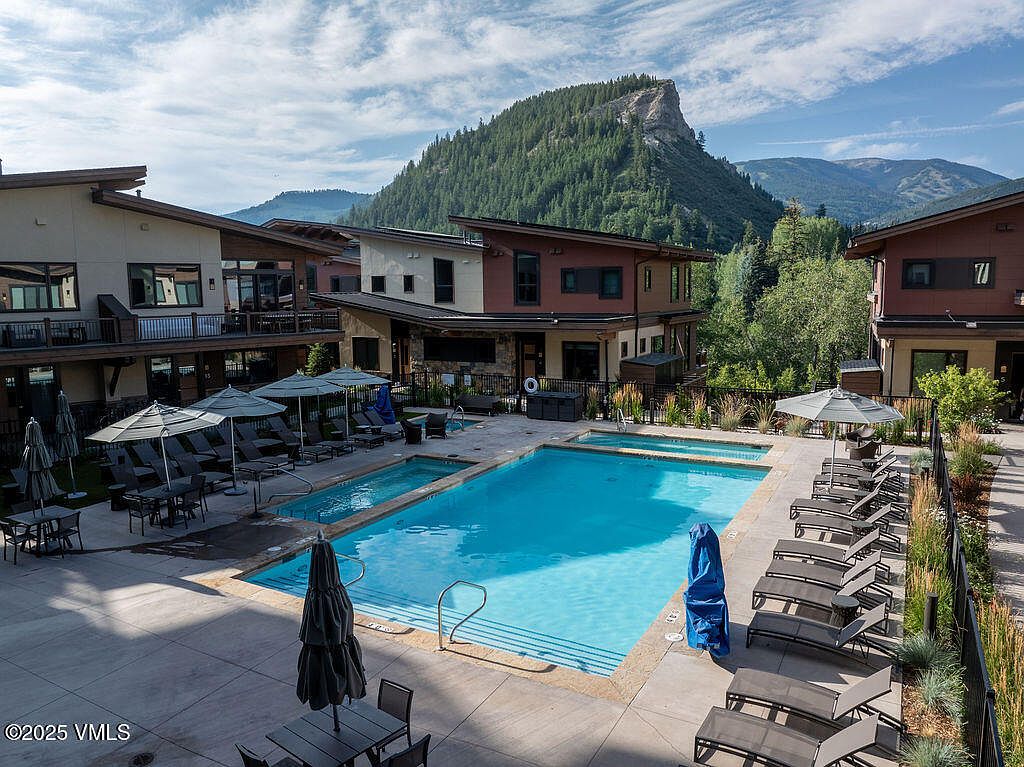 This image showcases a luxurious outdoor pool and spa area, surrounded by modern residential buildings. The pool features clear blue water and is flanked by lounge chairs and umbrellas, creating an inviting space for relaxation. The backdrop includes a scenic mountain view, enhancing the property's appeal and offering a sense of tranquility.