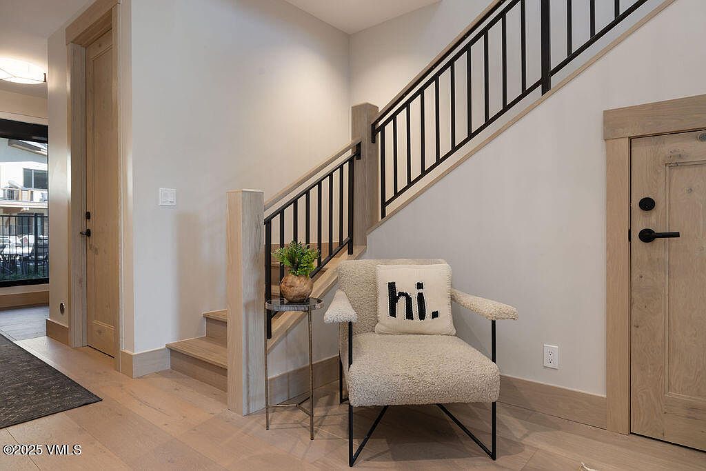 This interior shot showcases a modern hallway with a staircase. The stairs feature light wood treads and black metal railings, complemented by light-colored walls and wood trim. An accent chair with a "hi." pillow adds a welcoming touch, and a small side table with a potted plant enhances the decor.