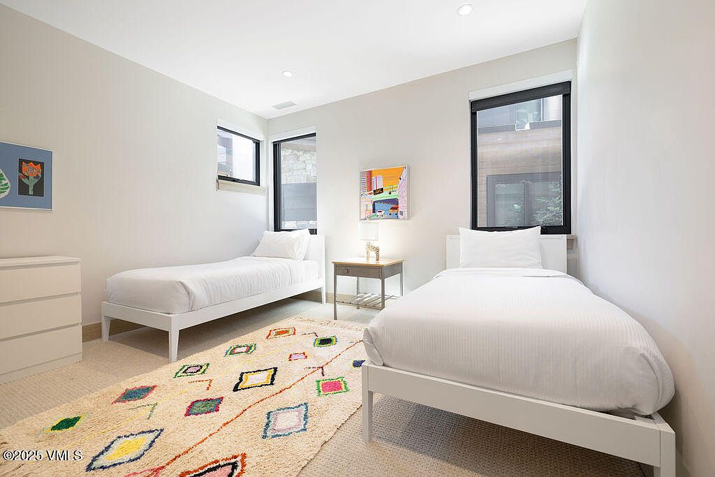 This is a bright and airy guest bedroom featuring two twin beds with white linens and frames. A colorful rug adds a touch of warmth to the neutral carpet, and natural light streams in through the windows. The room is simply decorated with artwork and a small side table, creating a clean and inviting space.