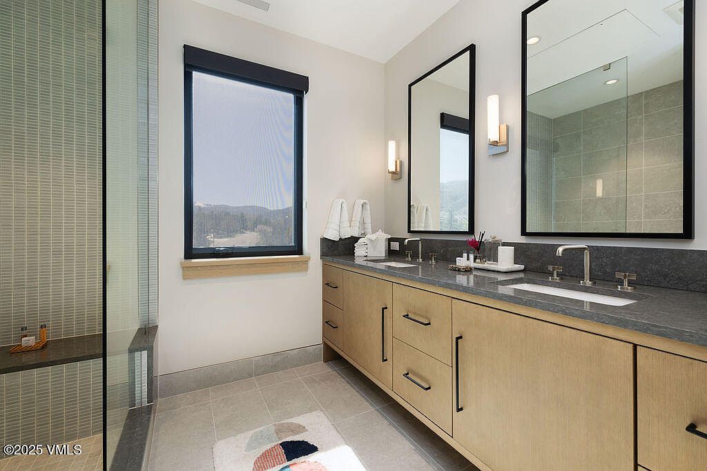 This is a well-lit primary bathroom featuring a double vanity with light wood cabinetry and dark countertops. Two large, black-framed mirrors hang above the sinks, complemented by modern sconces. A glass-enclosed shower is visible to the left, and a window provides natural light and a view of the outdoors.