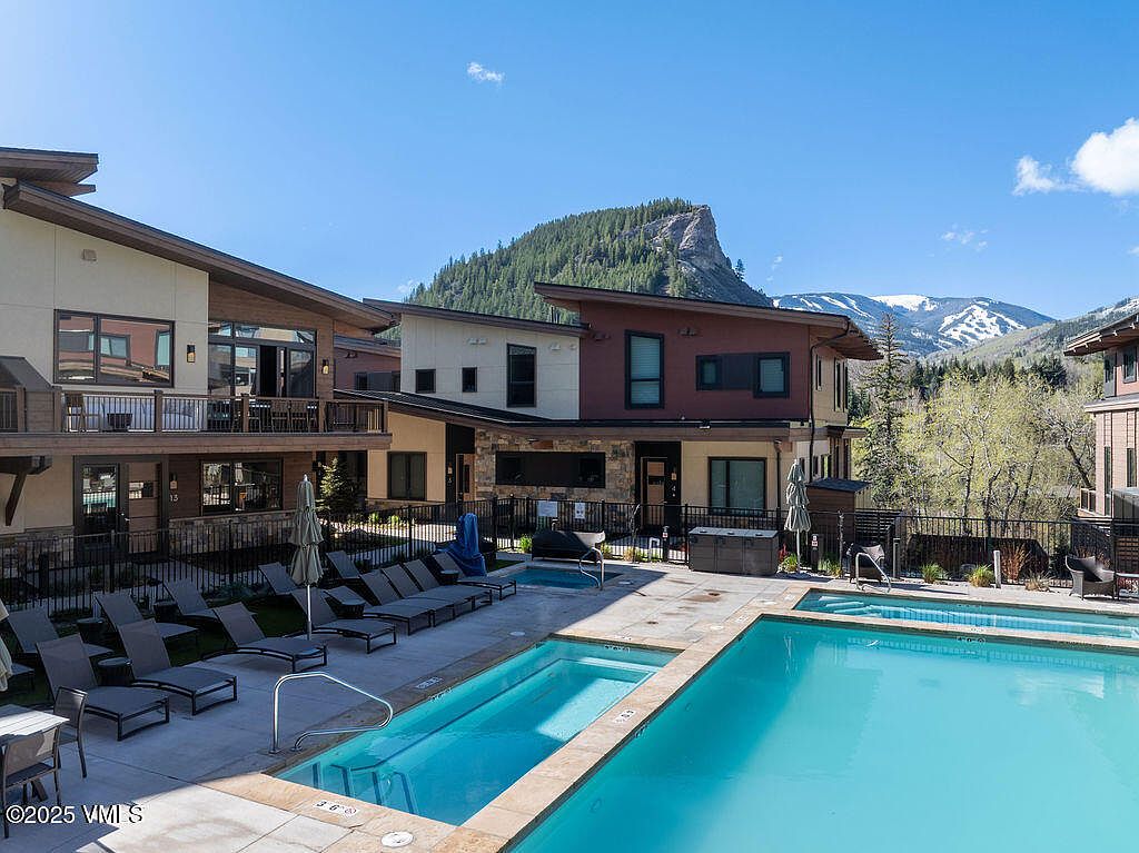 This image showcases a luxurious outdoor pool and spa area, surrounded by modern residential buildings. The pool features clear blue water and is lined with lounge chairs, creating an inviting space for relaxation. In the background, a scenic mountain view adds to the property's appeal, suggesting a serene and upscale living environment.