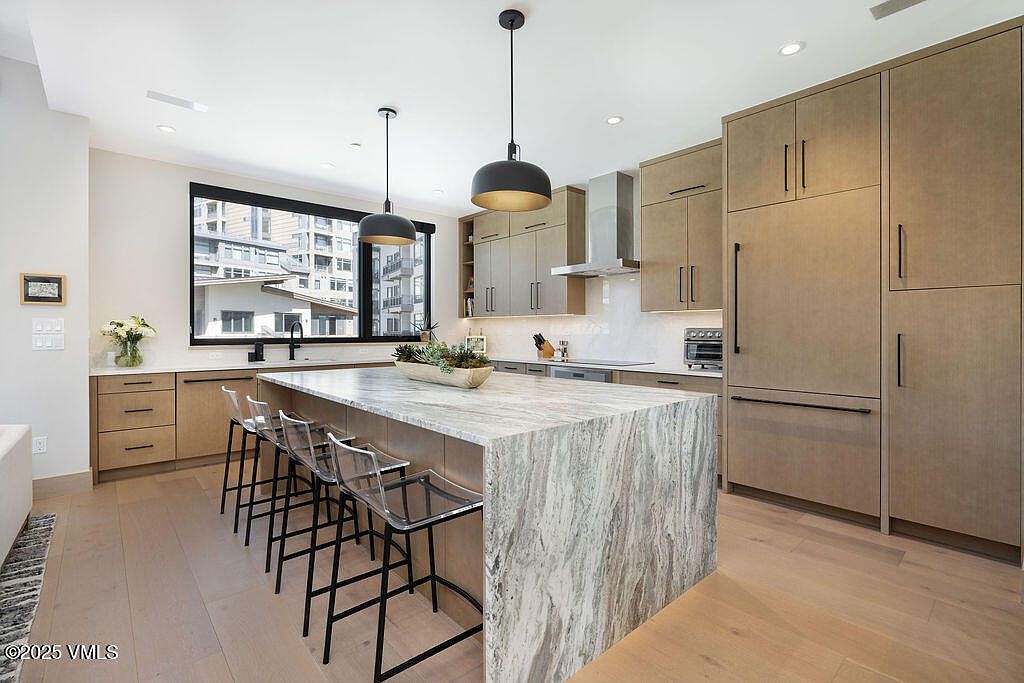 This is a well-lit, modern kitchen featuring light wood cabinetry and a large island with a striking marble countertop. The island is surrounded by clear bar stools with black frames, and pendant lights hang above. Stainless steel appliances and a large window complete the contemporary design, creating a clean and inviting space.